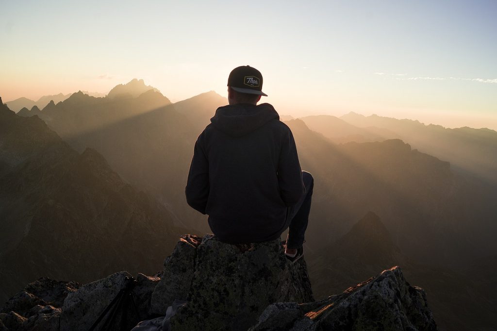 Symbolbild Handlungsaufforderung: Ein Mensch sitzt auf einem Berggipfel, entspannt auf einem Felsen. Der Blick ist auf die Weite des Gebirges gerichtet, wo die Sonne hinter den majestätischen Bergen auf- oder untergeht. Die Szene strahlt Ruhe und die Schönheit der Natur aus.