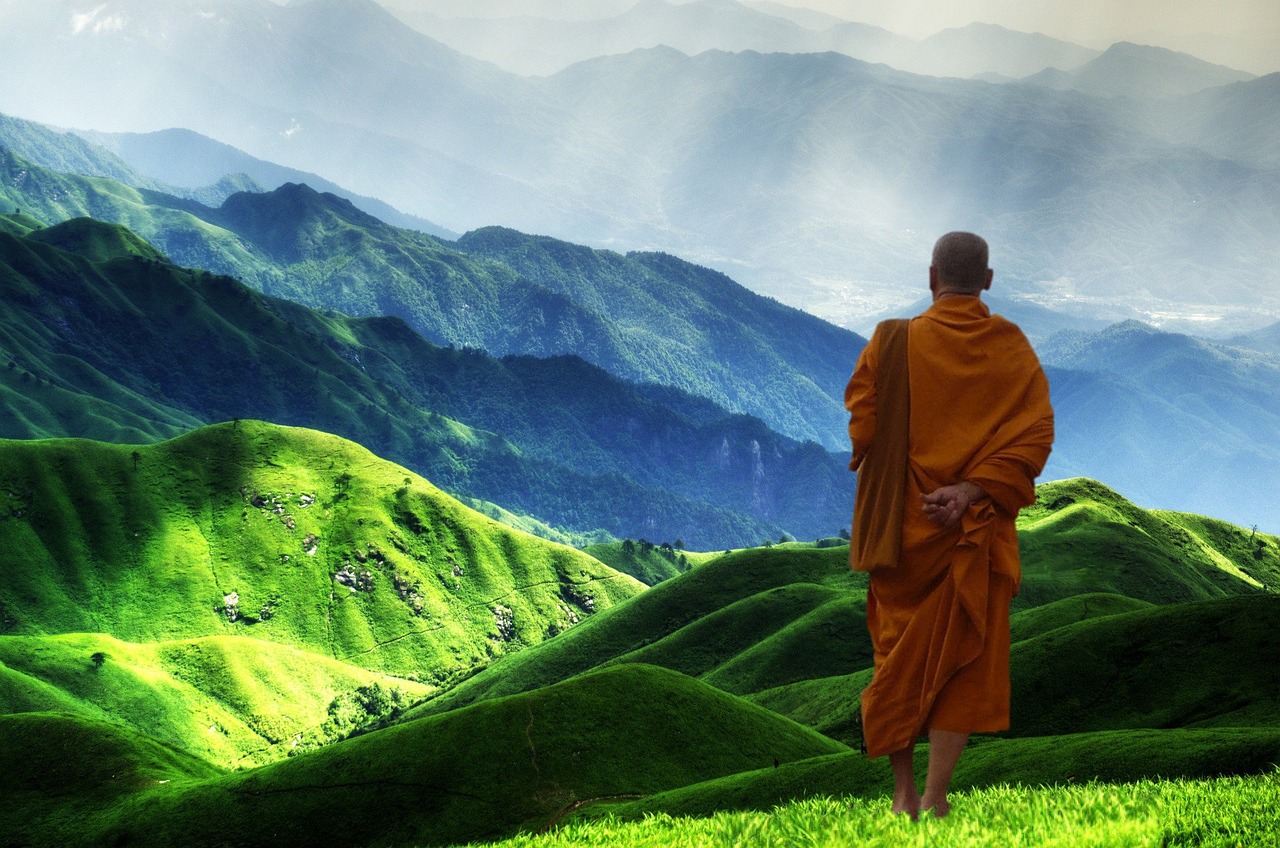 Hintergrundbild innere Stärke: Ein buddhistischer Mönch steht vor einem imposanten Gebirge und schaut in die Weite. Die Szene zeigt den Mönch von hinten, während er in stiller Kontemplation die majestätische Schönheit der Berglandschaft betrachtet.