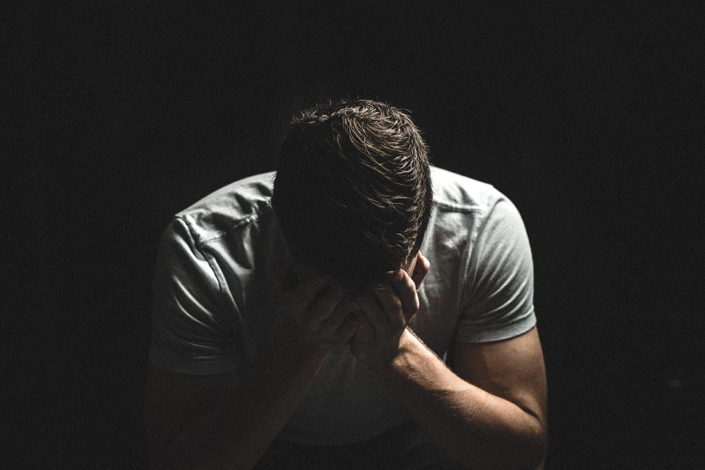 Symbolbild Stress: Ein Mensch schlägt die Hände vor dem Gesicht zusammen und neigt den Kopf. Das Bild ist von Dunkelheit umgeben, was die drückende Atmosphäre der Belastung und Anspannung betont.